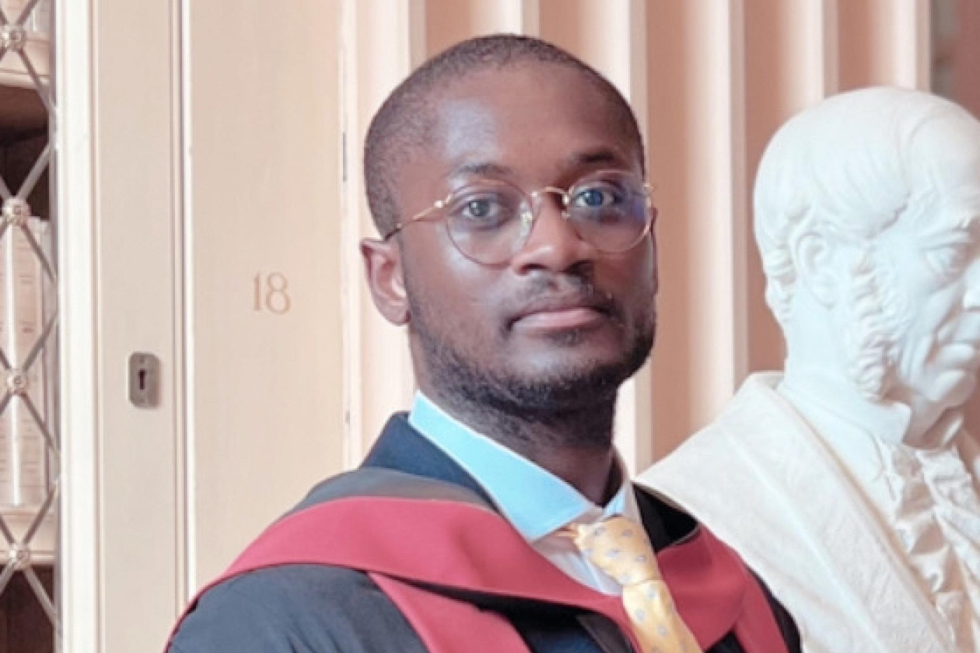 Kane wearing graduation robes, standing between books and a bust statue.