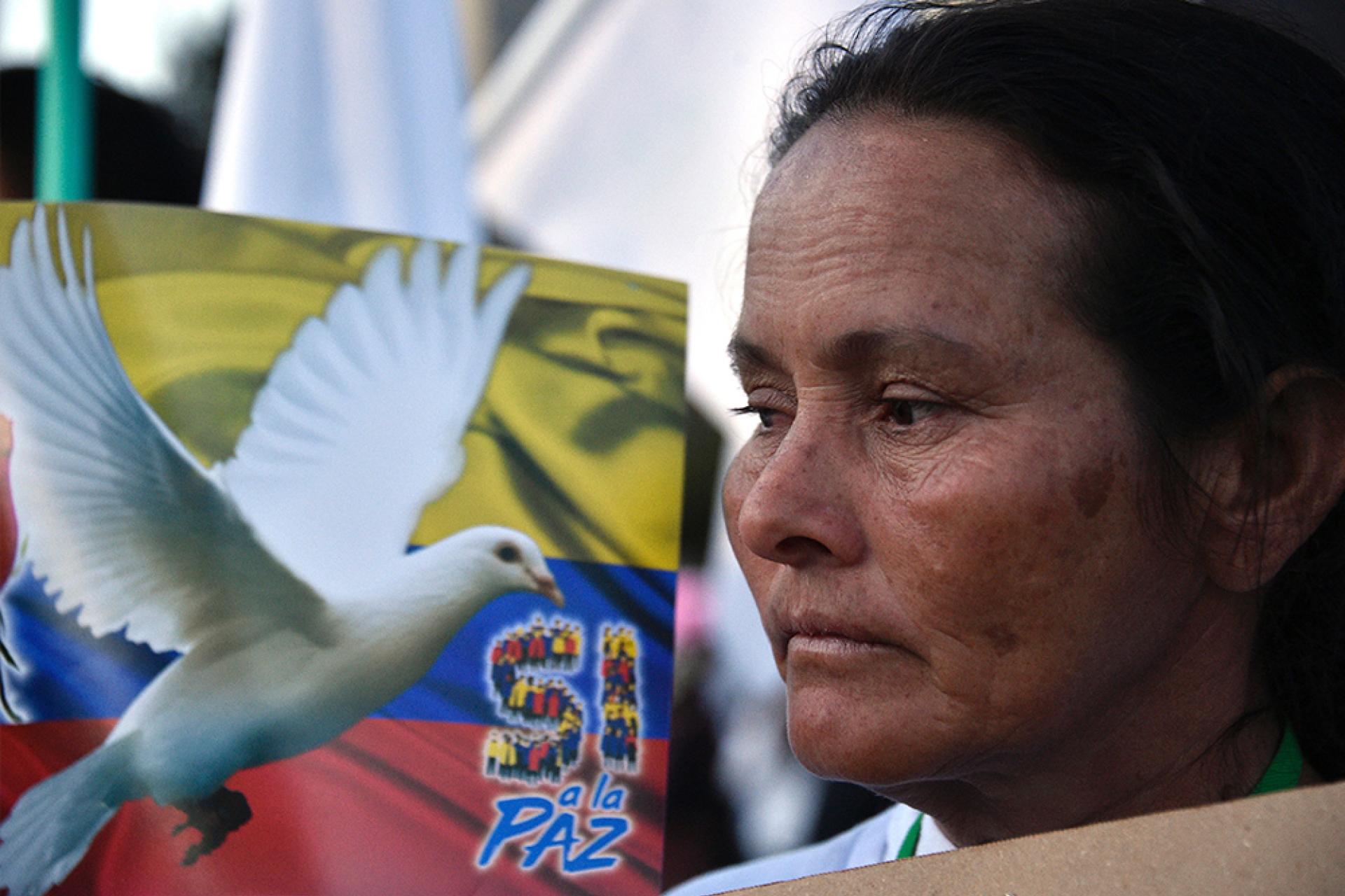 A woman stands in profile beside a poster showing a white dove and the words “Sí a la paz” (“Yes to peace”) against a Colombian flag background.