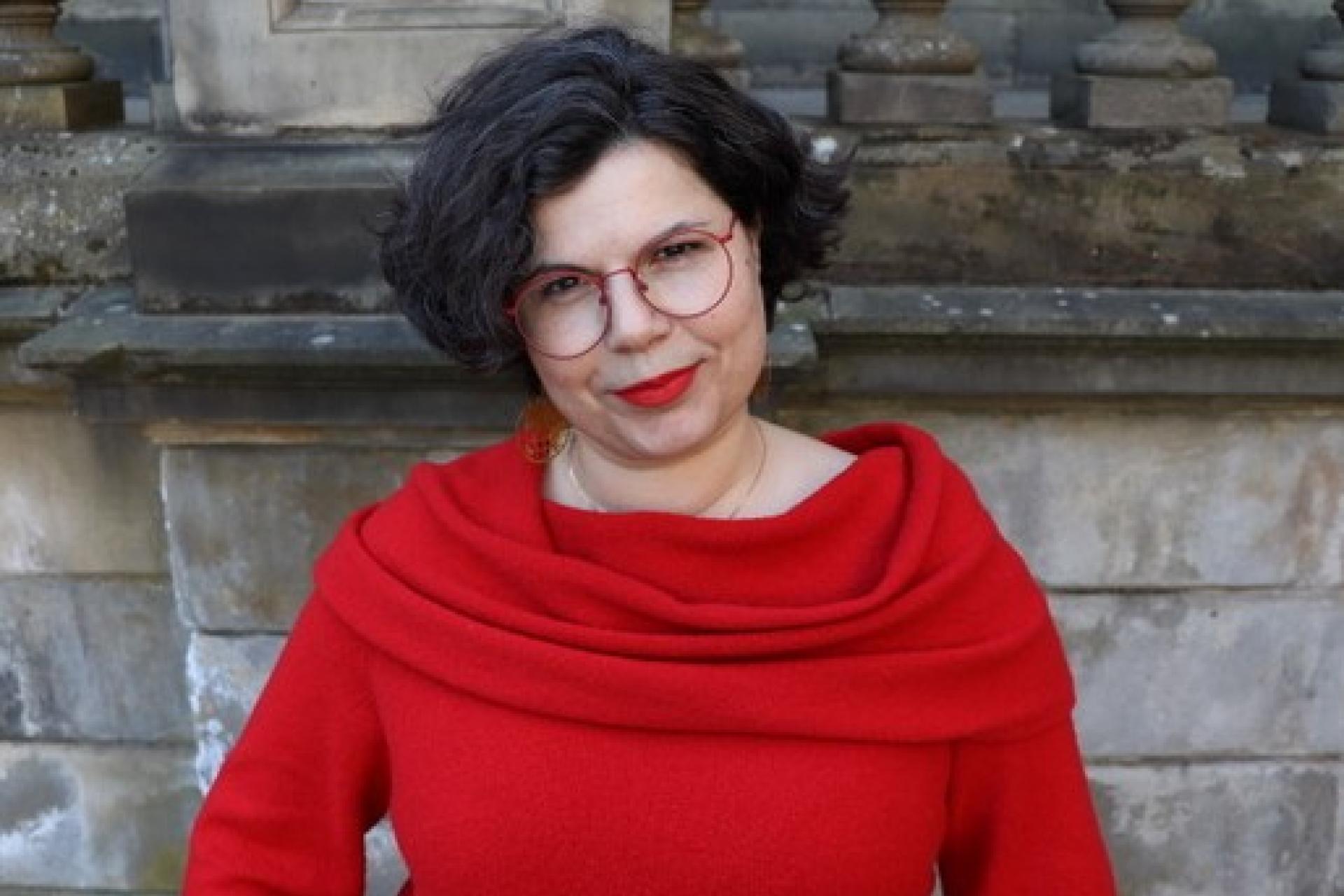 Dr Josephine Greenbrook smiles into the camera with a vibrant red jumper and glasses.
