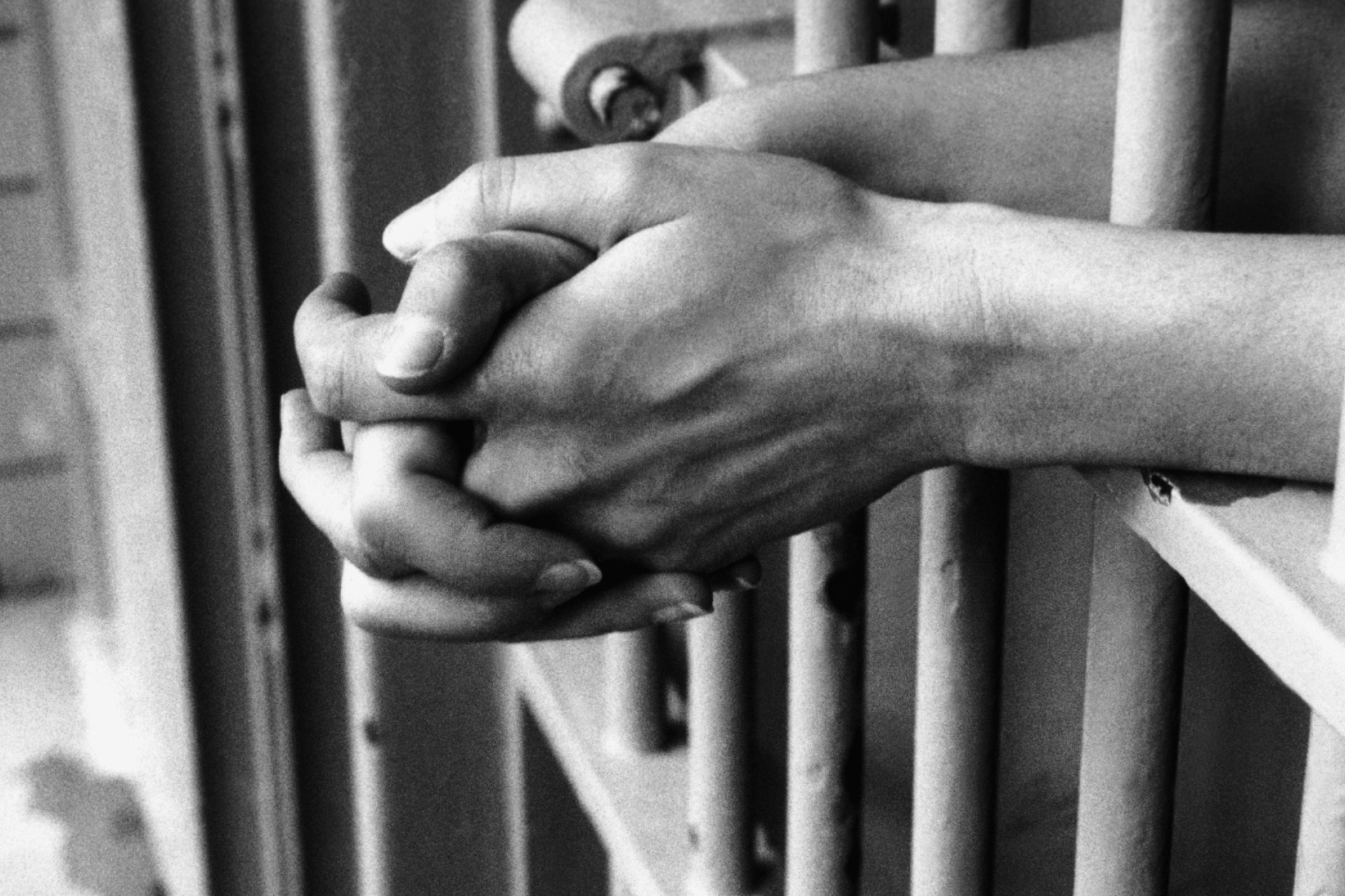 Hands clasped together rest through the bars of a prison cell in black and white. 